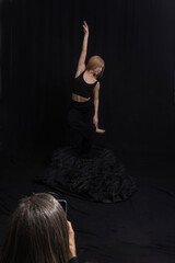 a dancer dressed in black, striking a dynamic pose against a dark background. One arm is raised elegantly above the head while the other extends downward.