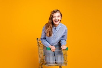 Young woman in casual attire posing playfully in a shopping cart on a vibrant yellow background