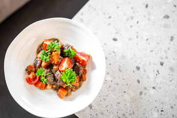Hearty homemade stew with meat chunks, vegetables, and cherry tomatoes, garnished with fresh parsley in a white bowl on a speckled countertop, close-up.