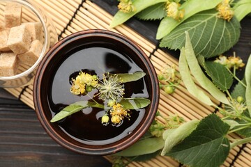 Fresh linden tea in cup, sugar cubes and flowers on dark wooden table, flat lay