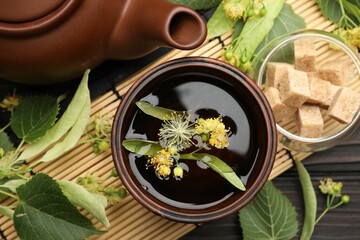 Fresh linden tea in cup, sugar cubes, teapot and flowers on dark wooden table, flat lay