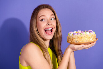 Excited young woman in casual attire holding a beautifully decorated cake against a purple...