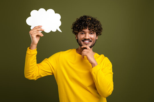 Young man holding a blank speech bubble displaying thoughtful and cheerful expression wearing a yellow shirt against solid background