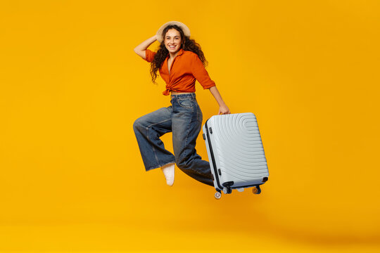 Cheerful lady traveler jumping with suitcase over orange background, woman in straw hat having fun, enjoying vacation time, full length, free space