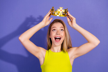 Cheerful young woman holding a golden crown with an excited expression, against a vibrant purple...