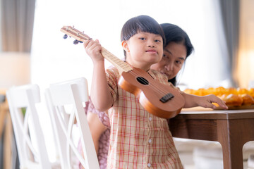 A child with Down syndrome playing ukulele while mother claps along, encouraging musical...