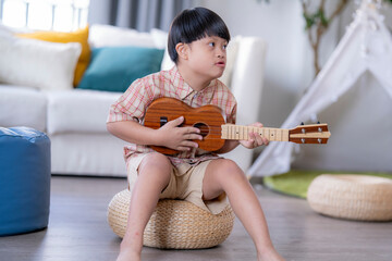Young child with Down syndrome concentrating while playing a ukulele, sitting indoors in a warm...