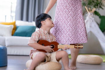 Young child with Down syndrome concentrating while playing a ukulele, sitting indoors in a warm...