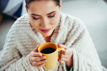 Charming Young woman enjoying warm coffee indoors, wrapped in blanket for comfort and recreation