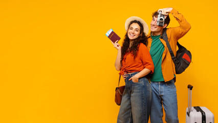 Cheerful tourists couple ready for vacation, posing with photo camera and luggage on yellow studio background, smiling at camera