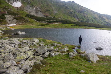 Outdoor enthusiast trekking across stone-covered lakeside in remote wilderness. Dramatic mountain scenery with cloudy sky and pristine water.