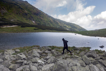 Hiker celebrating freedom by jumping across alpine brook, golden flowers framing scenic glacial valley with steep rocky slopes and snowfields