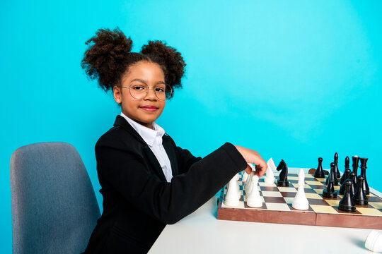 Confident young girl in formal attire playing chess on a turquoise background representing education and intellectual skills