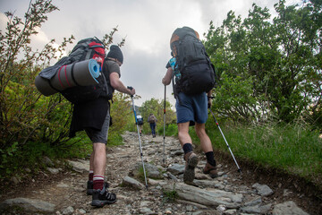 Two hikers with heavy backpacks and trekking poles climbing rocky mountain trail. Backpackers navigating challenging terrain on wilderness adventure.