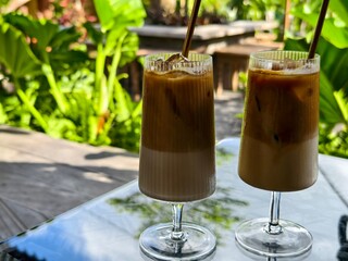 Two iced lattes served in tall glass goblets with eco-friendly straws in a tropical garden cafe