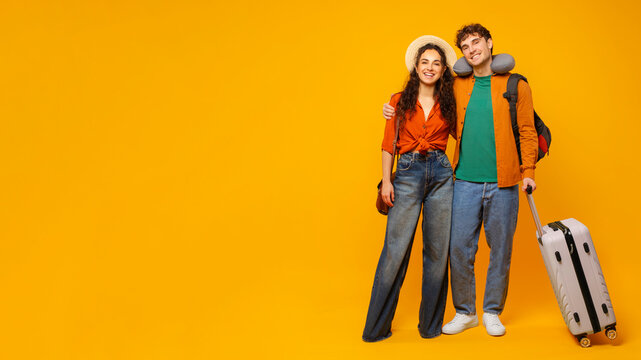 Happy man and woman getting ready for trip with suitcase, man wearing travel neck pillow and backpack, posing and embracing on yellow background - Powered by Adobe