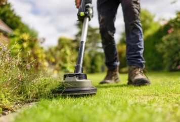 Individual operates an electric string trimmer in a residential backyard to neatly cut grass and control greenery overgrowth on a clear day