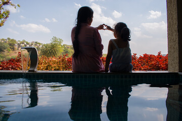 Indian Mother with her little daughter sitting near swimming pool in summer holiday. making heart with their hands.