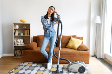 Tired woman with vacuum cleaner standing in modern living room, showing fatigue from domestic...