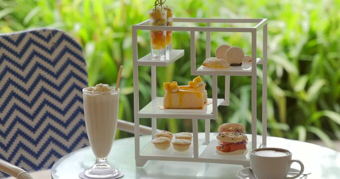 Afternoon tea set on elegant table in tropical garden, featuring refreshing milkshake, delectable desserts, macarons, and coffee. Cozy, relaxing outdoor cafe ambiance.