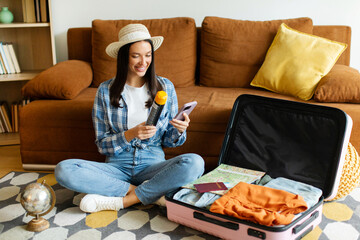 Woman preparing for vacation, packing clothes in suitcase and checking list on phone, sitting near...