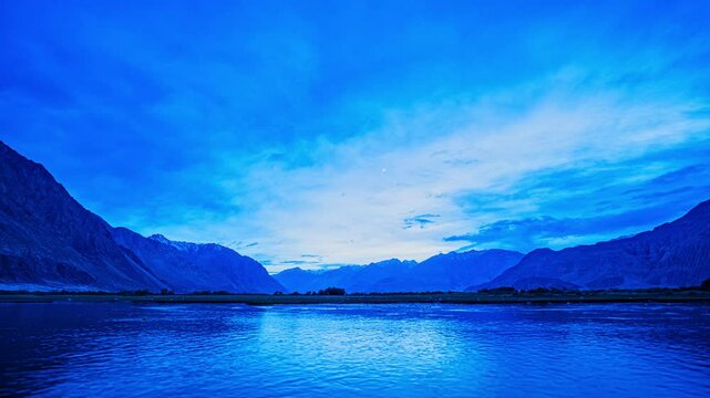 timelapse of Himalayas and blue sky mirrored in the Shyok River at sunrise, near lush Sumur village&mdash;where green orchards and barley fields thrive against Nubra Valleys stark mountain beauty.