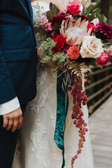 Close up of bride and groom holding vibrant cascading wedding bouquet with king protea, roses, and greenery, bride in lace dress and groom in navy suit, romantic wedding floral detail photo © Jordan