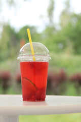 Lemonade in a disposable plastic cup, takeaway drink. Refreshing drink against the background of a park in summer. Pink lemonade with raspberries and lime. Close-up of a drink in a disposable cup