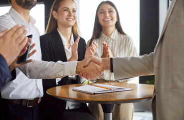 Obraz premium Two business professionals shaking hands, making agreement or deal in office. Smiling colleagues standing by table with documents, clapping hands and celebrating successful partnership and teamwork.