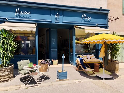 Gruissan, France - May 30, 2025: Charming storefront of a boutique named Maison de Plage featuring yellow striped parasol outdoor seating and lush greenery creating a welcoming atmosphere for shoppers