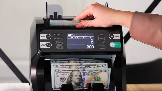 Hand placing US dollars into an electronic money counter. A close-up view of a person inserting a stack of US dollar bills into a digital currency counting machine.