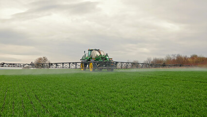 Fototapeta premium Agricultural sprayer tractor working on green crop field. A large green and yellow agricultural sprayer sprays water over a lush green field under a cloudy sky.