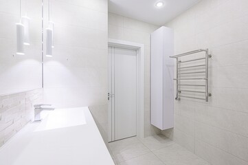 bright, minimalist bathroom featuring white tiles, a sleek countertop with a modern faucet, a towel warmer, and cylindrical pendant lights