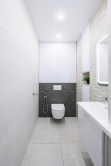 Modern, narrow bathroom with white and gray tiles, sleek white cabinets, a wall-mounted toilet, and recessed lighting
