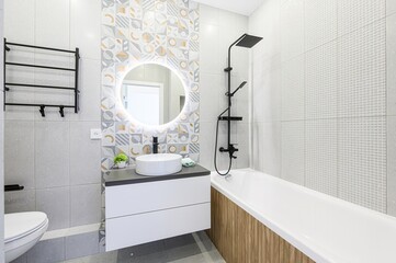 Modern bathroom with white suite, round lighted mirror, black fixtures, and stylish patterned & textured tiles