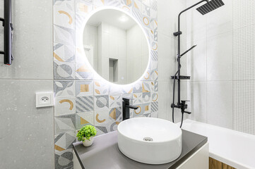 Modern bathroom with white suite, round lighted mirror, black fixtures, and stylish patterned &...