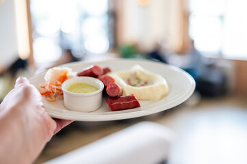 Close-up of a hand holding a white plate with mashed potatoes, sliced sausages, green sauce, and pickled vegetables. Blurred indoor background, casual dining concept.