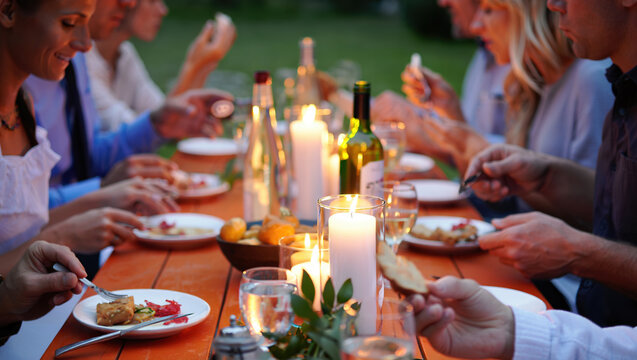 close-up of an outdoor dinner, a table covered with a white tablecloth, with various snacks, glasses, bottles and lit candles. The hands of people sitting at the table are visible. - Powered by Adobe