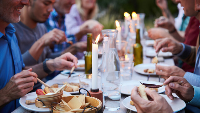 close-up of an outdoor dinner, a table covered with a white tablecloth, with various snacks, glasses, bottles and lit candles. The hands of people sitting at the table are visible. - Powered by Adobe