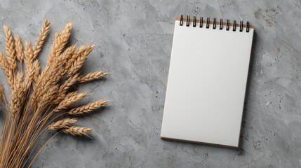A blank notepad rests on a smooth grey surface, accompanied by a cluster of dried wheat. This setup provides a serene and minimalistic aesthetic for creative expression or planning