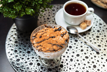Tiramisu dessert with cocoa and coffee cup. Classic Italian tiramisu served in a glass dish, topped with cocoa powder and chocolate drizzle, paired with hot black coffee.
