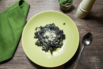 Squid Ink Risotto with Parmesan on Green Plate. Dark squid ink risotto garnished with grated Parmesan cheese, served on a green plate over a rustic wooden table setting.