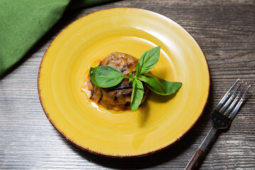 Baked Eggplant Parmigiana with Basil on Yellow Plate. Elegant serving of baked eggplant parmigiana topped with fresh basil, plated on a rustic yellow dish over wooden table surface.