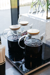 Glass Jugs of Herbal Iced Tea with Wooden Lids. Transparent glass teapots filled with dark herbal iced tea, sealed with wooden lids, displayed on a countertop in a bright setting.