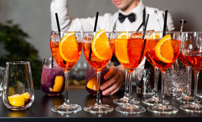 Orange cocktails on bar counter with bartender. Row of vibrant orange cocktails with orange slices and straws on a bar counter, with bartender preparing drinks in background.