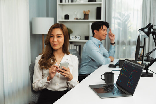 Profile smiling beautiful Asian businesswoman looking at camera, using phone with laptop at modern office on working desk casual day. Blurry background man colleague analyzing data on pc. Infobahn.