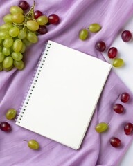 Arranging red and green grapes around a white notebook on soft purple cloth a creative still life composition for photography inspiration.