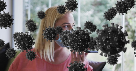 Woman wearing mask, salmon top, speaking into smartphone on sofa by window, with 3D virus particles