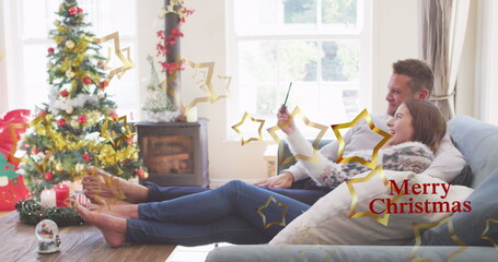 Reclining couple in sweaters taking selfie in Christmas living room, with tree, stove, copy space