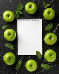 Captivating arrangement of green apples and fresh mint leaves surrounding a white notepad on luxurious black marble surface in a minimalist flat lay style.